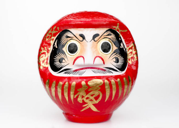 Round red doll with a white inset featuring a face and some butterfly-like patterns and some golden patterns and Japanese text in the bottom and on the sides