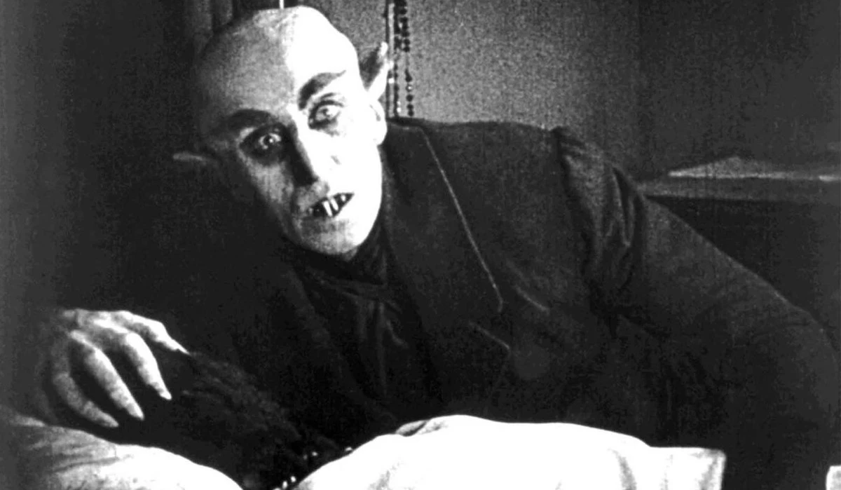 Black-and-white still of a bald Max Schreck with prominent teeth, grasping a black mass behind a bed